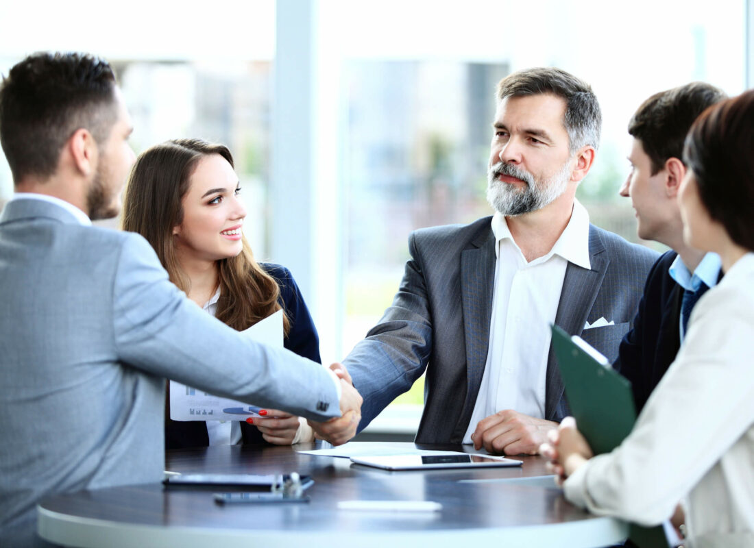 A group of business professionals sitting at a table. Two of which are shaking hands over documents.