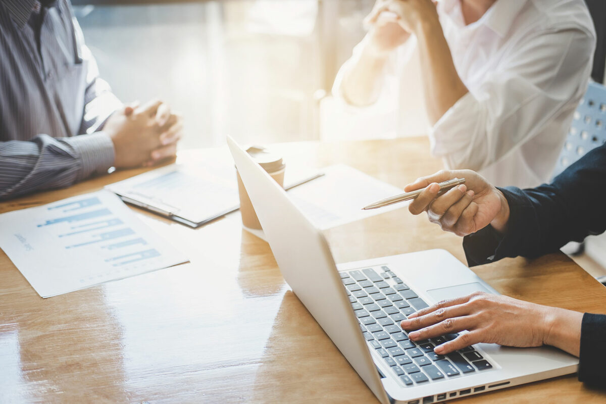 Three business professionals sitting at a table discussing and pointing to computer and papers.